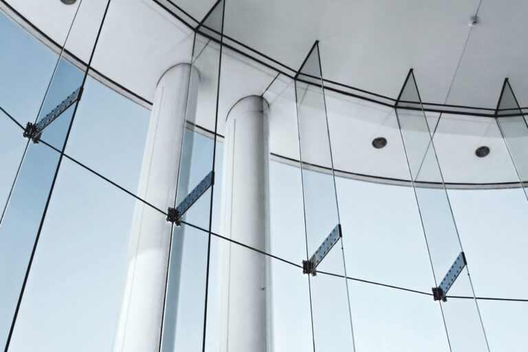 Das Bild zeigt eine moderne Architektur mit großen Glasflächen, die von Metallhalterungen und Seilen getragen werden. Die Glasstruktur ist gebogen und wird von weißen Säulen sowie einer Decke mit eingelassenen Leuchten ergänzt. Durch die Glasflächen ist der Himmel sichtbar, was auf einen Außenbereich oder ein Atrium hinweist.