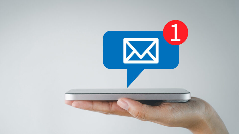 Hand mit Smartphone, über dem ein Benachrichtigungssymbol in Form einer blauen Sprechblase mit Briefumschlag und rotem Kreis mit der Zahl „1“ schwebt, was auf eine neue Nachricht hinweist.