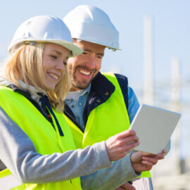 Zwei Personen mit weißen Schutzhelmen und gelben Warnwesten im Außenbereich, eine davon hält ein Tablet, beide blicken darauf.