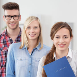 Drei Personen stehen nebeneinander, die rechte Person hält eine blaue Mappe mit der Aufschrift „BEWERBUNG“.