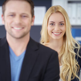 Zwei Personen stehen nebeneinander, die linke trägt ein dunkles Sakko über einem blauen Hemd, die rechte hat lange blonde Haare und trägt ein dunkles Oberteil; im Hintergrund befinden sich Regale mit Büchern und Ordnern.