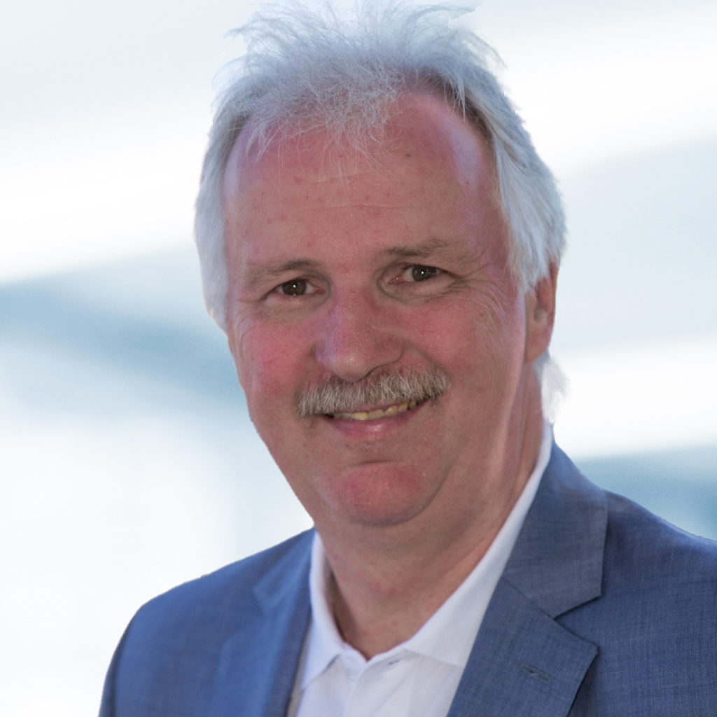 Thomas Karg trägt ein blaues Sakko und ein weißes Hemd. Der Hintergrund ist unscharf und zeigt ein Büro.