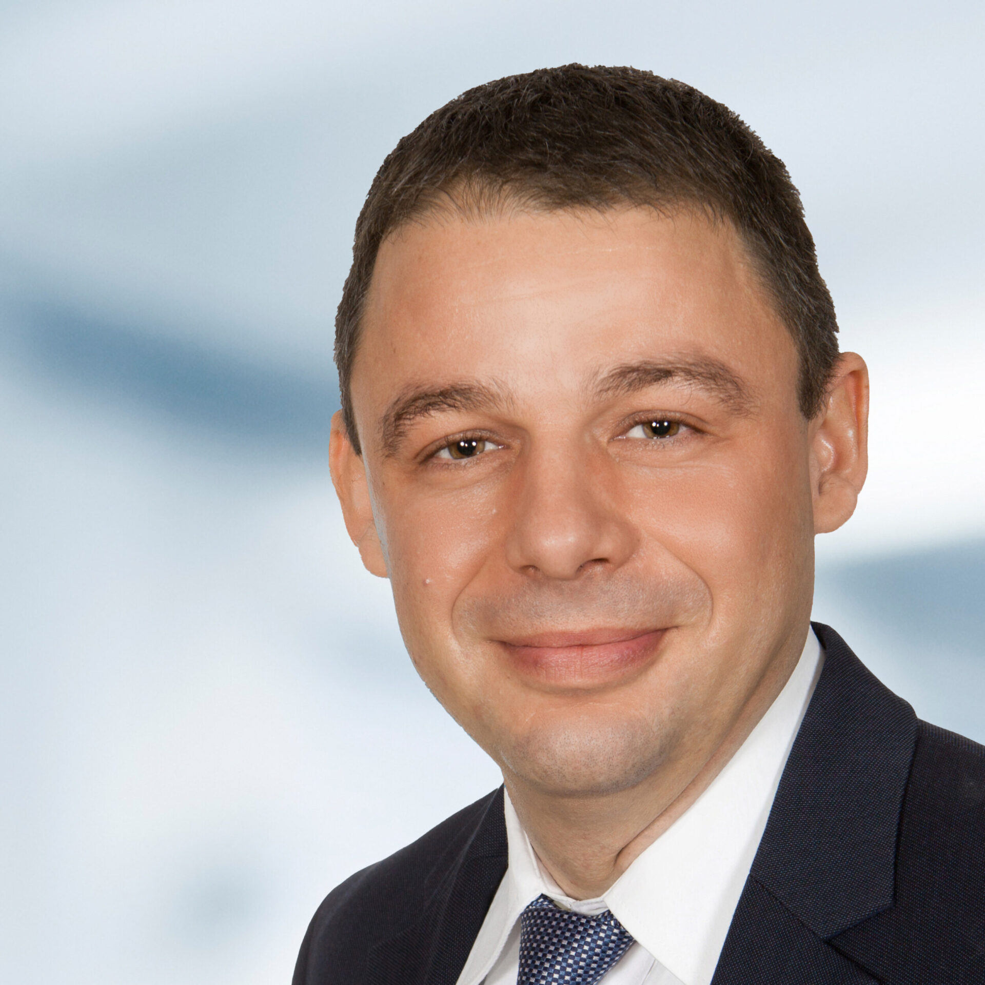Andreas Klug trägt einen Anzug mit Krawatte. Der Hintergrund ist unscharf und zeigt ein Büro