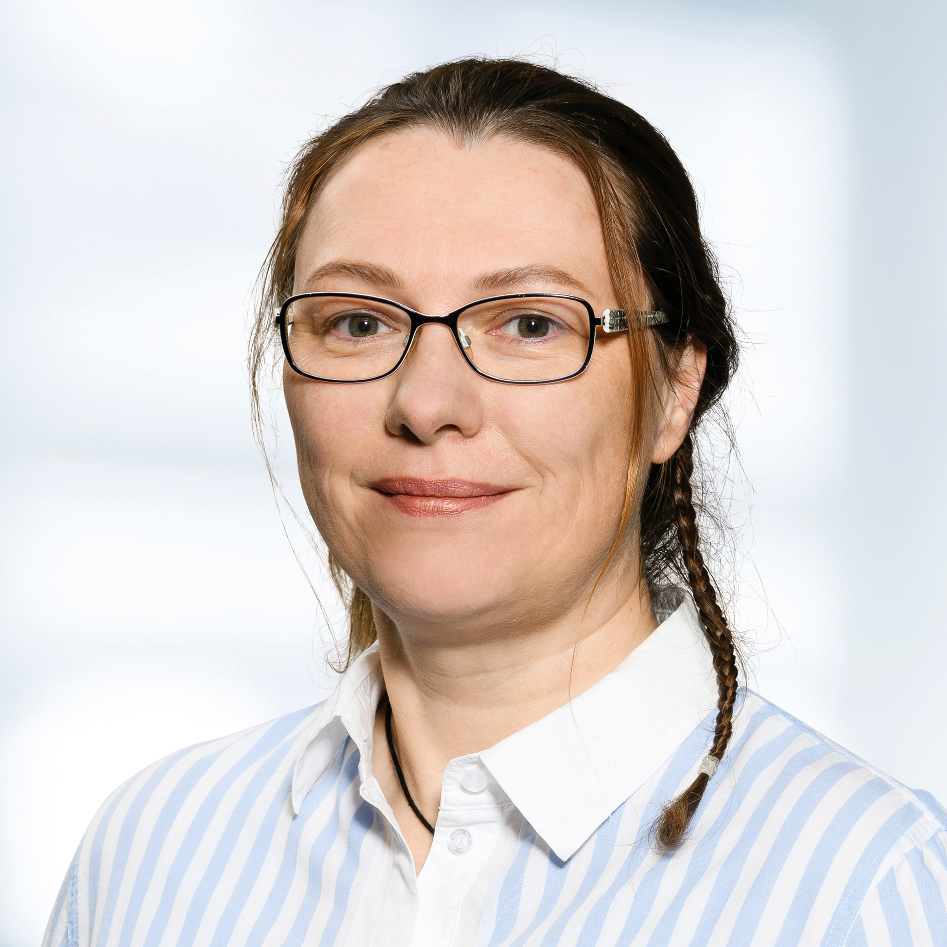 Barbara Koch trägt eine Brille und ein blau-weiß gestreiftes Hemd. Der Hintergrund ist unscharf und zeigt ein Büro.