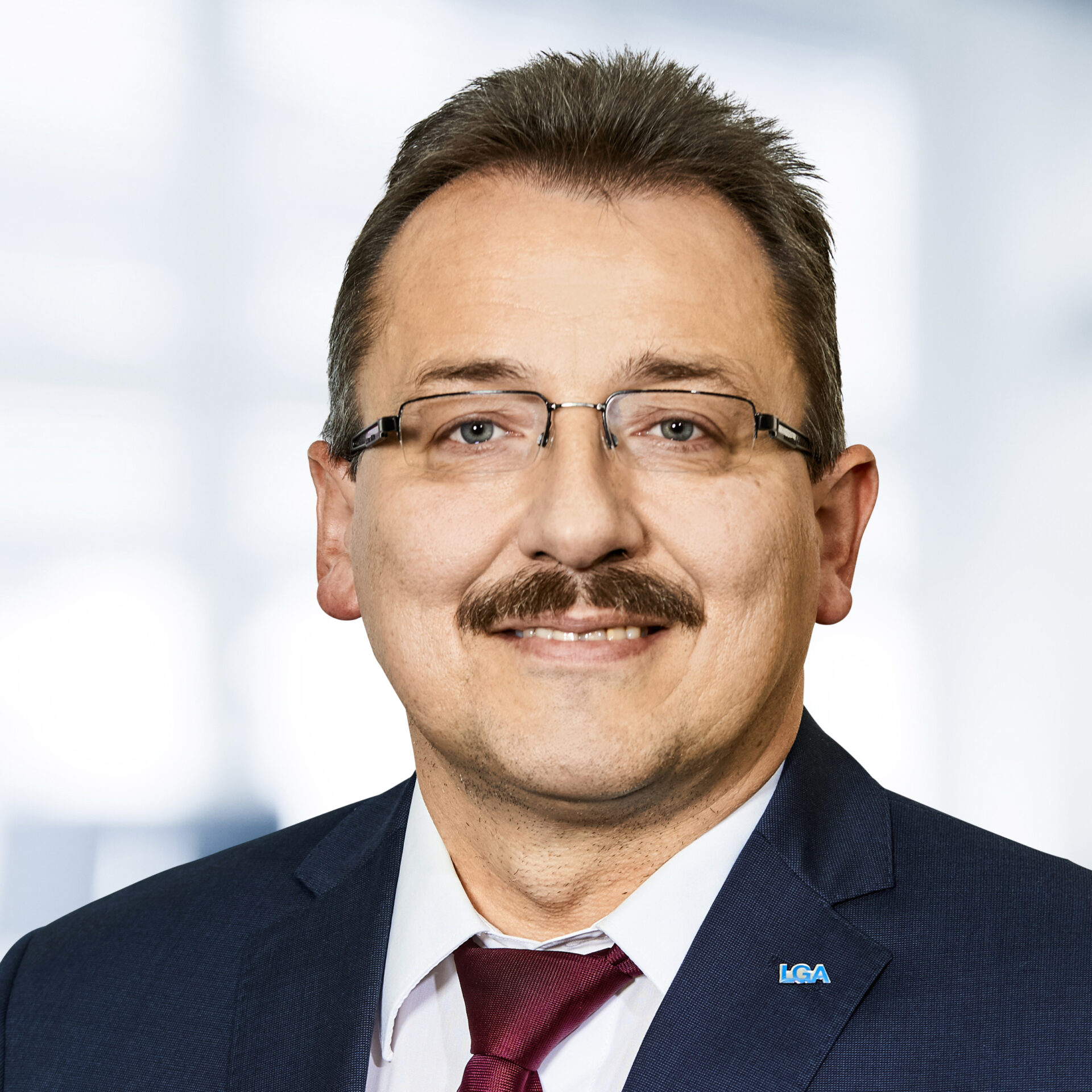 Martin Koller trägt einen dunklen Anzug, ein weißes Hemd und eine rote Krawatte. Der Hintergrund ist unscharf und zeigt ein Büro.