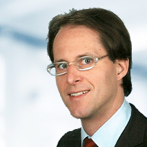 Simon Kröniger trägt eine Brille, ein hellblaues Hemd, ein dunkles Sakko und eine rote Krawatte. Der Hintergrund ist unscharf und zeigt ein Büro.