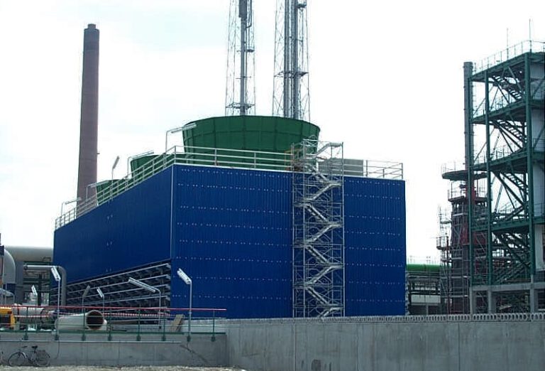 Das Bild zeigt eine industrielle Anlage mit einem großen blauen Gebäude, dessen oberer Bereich grün gestaltet ist. An der Seite des Gebäudes befindet sich ein Gerüst, und im Hintergrund sind hohe Schornsteine sowie Metallkonstruktionen zu sehen. Es handelt sich vermutlich um einen Teil eines Kraftwerks oder eines Industriekomplexes.