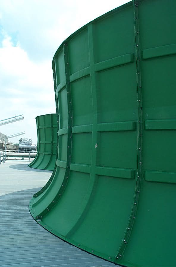 Das Bild zeigt große, grüne, gebogene Industrieanlagen oder Tanks mit horizontalen und vertikalen Verstärkungen. Sie stehen auf einer grauen Plattform mit Geländern. Im Hintergrund sind weitere industrielle Elemente wie Kräne und Ausrüstungen zu sehen, der Himmel ist teilweise bewölkt.