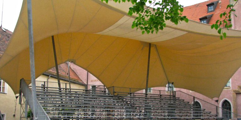 Das Bild zeigt eine überdachte Sitztribüne im Freien mit Metallbänken. Ein großes, beigefarbenes Stoffdach, das von mehreren Masten getragen wird, spendet Schatten über dem gesamten Bereich. Im Hintergrund sind Gebäude mit roten Ziegeldächern sowie etwas Grün im Vordergrund zu sehen.