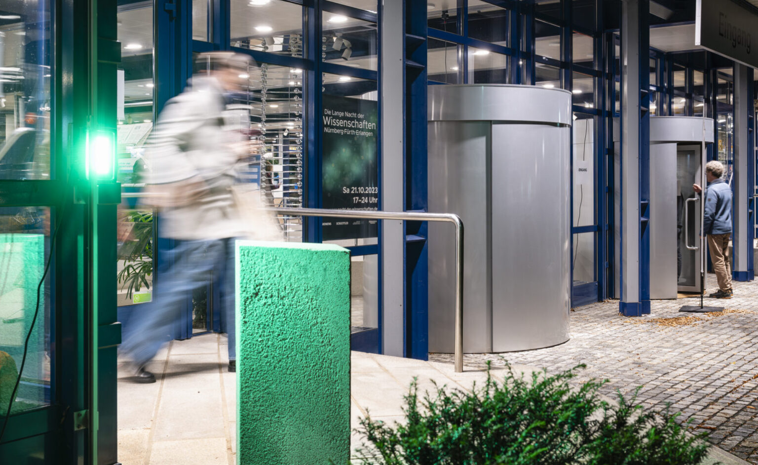 Eine Person betritt durch einen Eingang mit grünem Licht eine Veranstaltung; im Hintergrund ist ein Plakat zur „Langen Nacht der Wissenschaften“ am 21.10.2023 in Nürnberg, Fürth und Erlangen zu sehen.