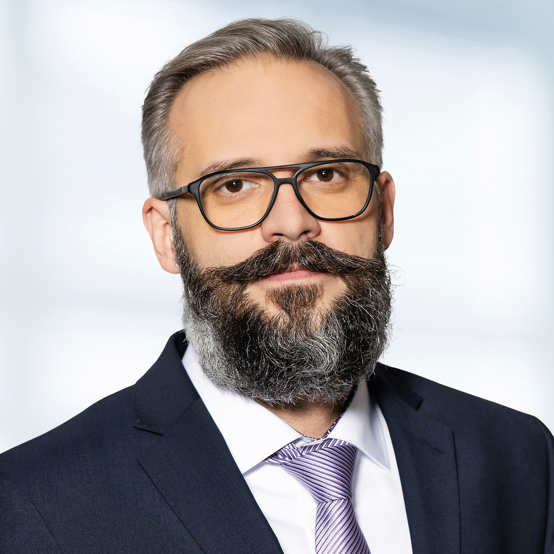 Jürgen Lehnert trägt einen dunklen Anzug, ein weißes Hemd und eine helllila Krawatte. Der Hintergrund ist unscharf und zeigt ein Büro.