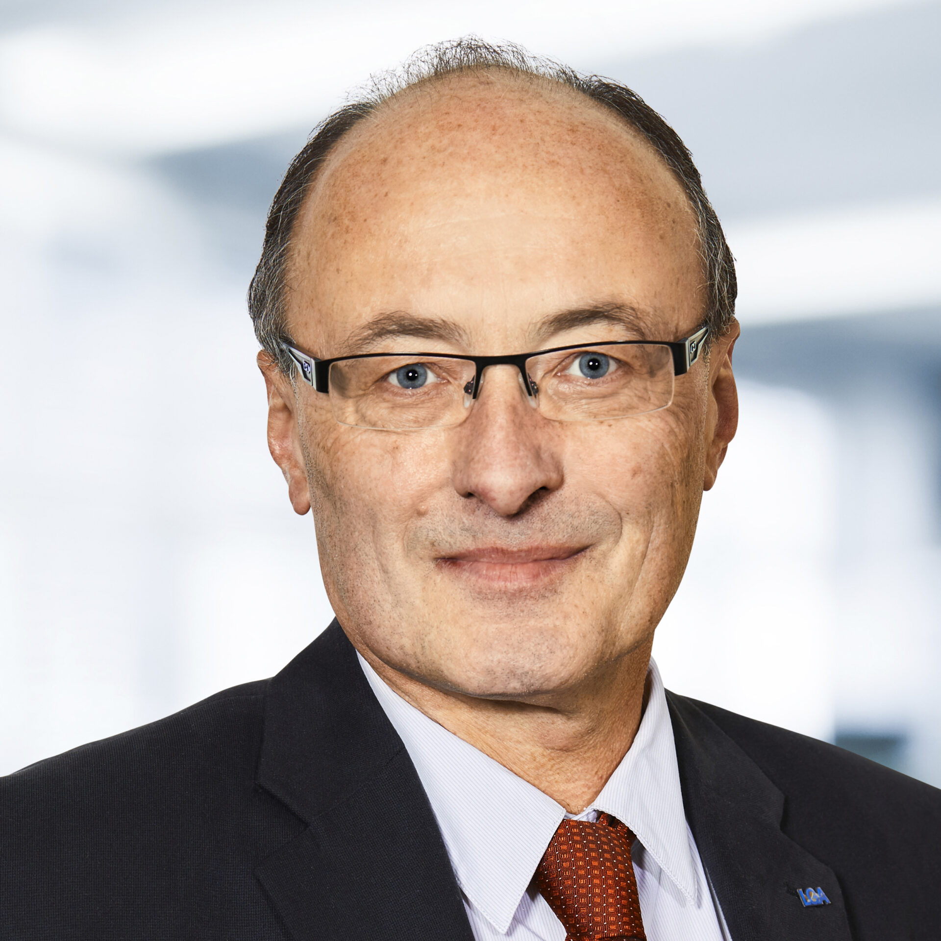 Dr. Ulrich Leißner trägt einen dunklen Anzug, ein weißes Hemd und eine rote Krawatte. Der Hintergrund ist unscharf und zeigt ein Büro.