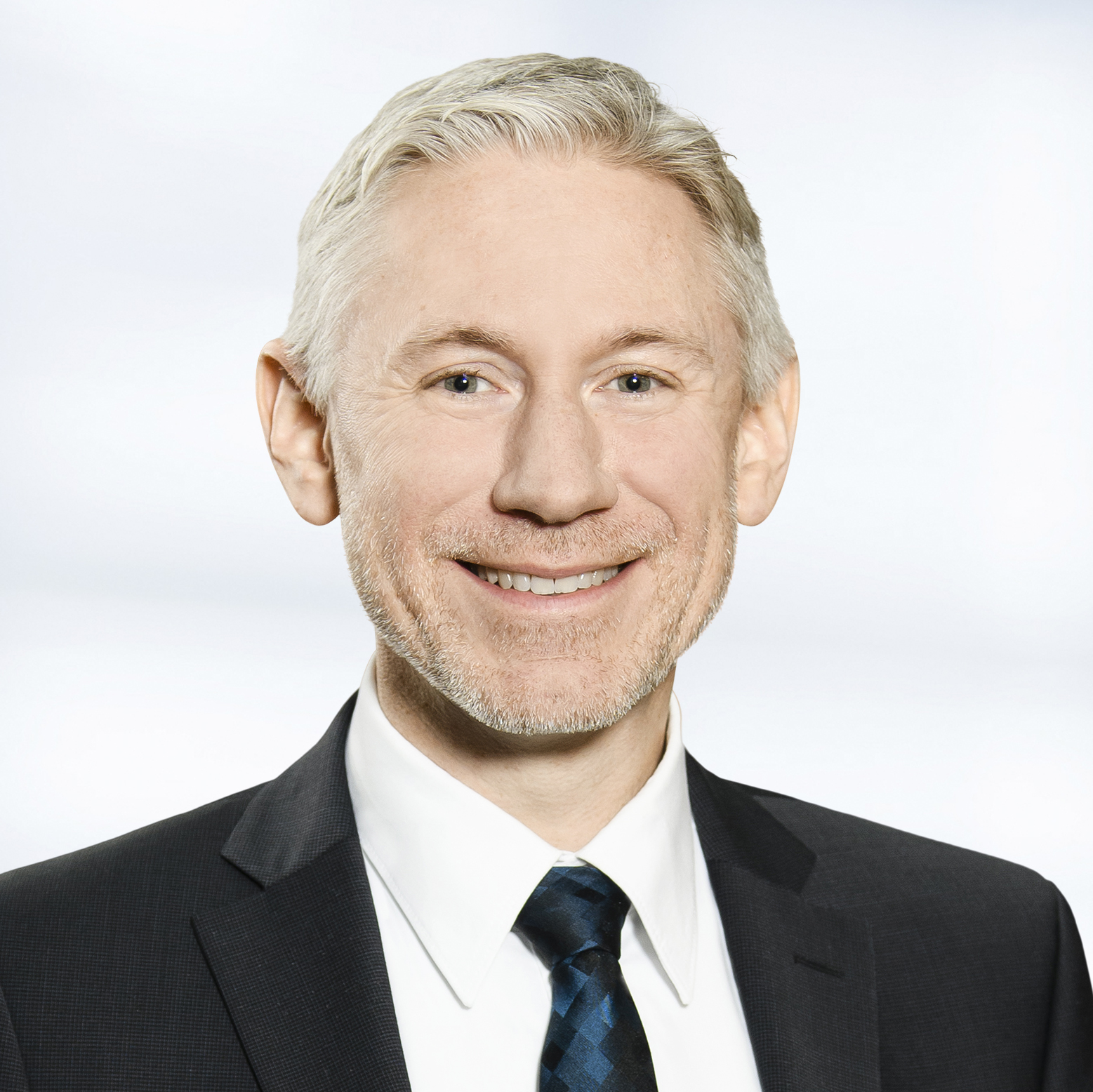 Stefan Lenhart trägt einen dunklen Anzug, ein weißes Hemd und eine blau gemusterte Krawatte. Der Hintergrund ist unscharf und zeigt ein Büro.