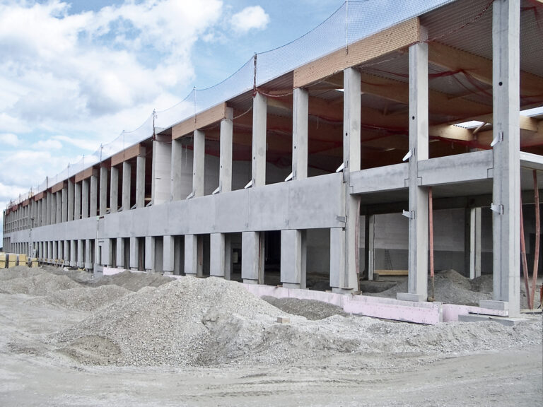 Das Bild zeigt ein großes Gebäude im Rohbau mit mehreren Betonstützen und Trägern. Im Vordergrund liegen Schotter- und Erdhaufen, am oberen Rand des Baus ist ein Sicherheitsnetz angebracht.