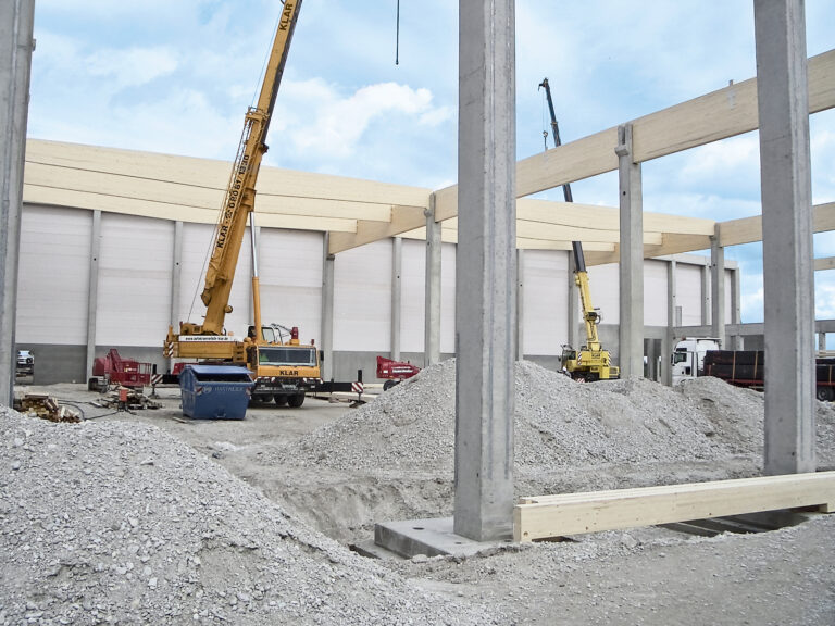 Das Bild zeigt eine Baustelle mit mehreren großen Betonpfeilern und Trägern sowie zwei Kränen – einem gelben und einem orangefarbenen. Im Hintergrund steht ein teilweise errichtetes Gebäude mit heller Fassade und offenen Wandflächen.