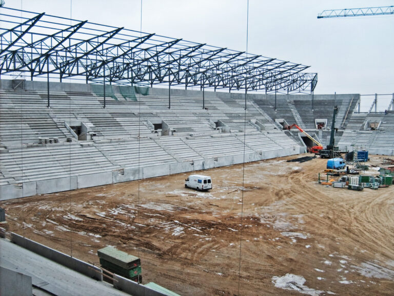 Das Bild zeigt eine Stadion-Baustelle mit teilweise fertiggestellten Betonsitzreihen und einem Stahlgerüst für das Dach. Auf dem erdigen Boden befinden sich Baumaterialien, Maschinen und ein weißer Transporter, im Hintergrund steht ein Kran.