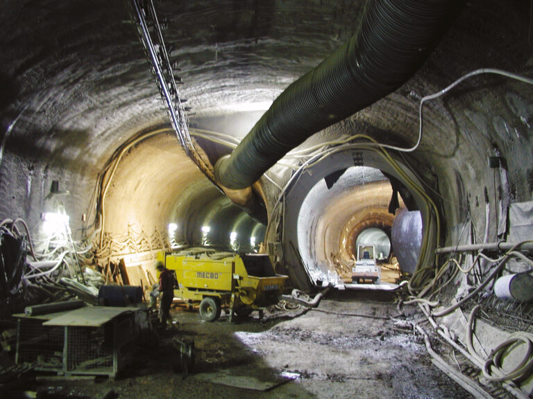 Das Bild zeigt eine Baustelle im Inneren eines großen Tunnels mit mehreren Baumaschinen, darunter ein gelbes Fahrzeug. Entlang der Tunnelwände verlaufen Beleuchtung, Kabel und Schläuche, an der Decke ist ein großes Belüftungsrohr angebracht.