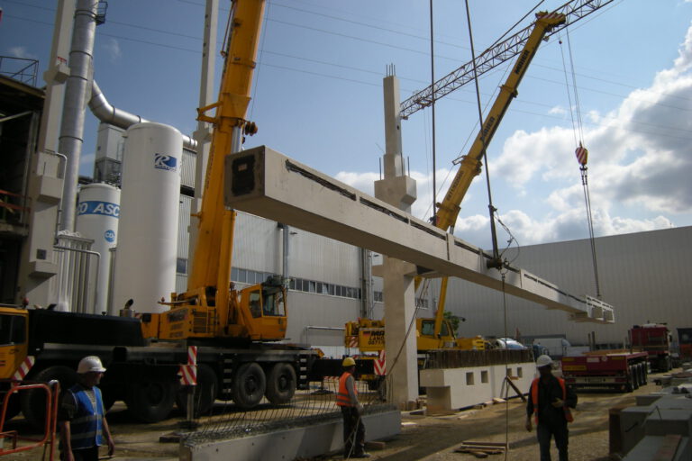 Das Bild zeigt eine Baustelle, auf der zwei große gelbe Kräne einen langen Betonträger anheben und ausrichten. Mehrere Arbeiter mit Schutzhelmen und Warnwesten beobachten den Vorgang, im Hintergrund sind Industrieanlagen und ein Gebäude zu sehen.