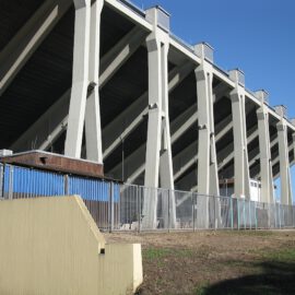 Das Bild zeigt die Außenansicht eines großen Stadions mit mehreren Betonstützen und Trägern. Hohe, senkrechte Pfeiler tragen den oberen Tribünenbereich, im unteren Bereich ist ein Metallzaun mit blauen Paneelen dahinter zu sehen.