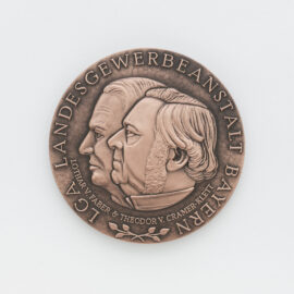 Bronzene Medaille auf weißem Hintergrund mit dem geprägten Profil zweier Köpfe in der Mitte, umgeben von der Inschrift „LGA LANDESGEWERBEANSTALT BAYERN“ und „LOTHAR V. FABER & THEODOR V. CRAMER-KLETT“.