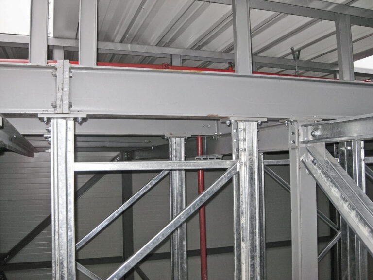 Das Bild zeigt einen Ausschnitt einer Metallkonstruktion mit Stahlträgern, Stützen und diagonalen Verstrebungen. Die Decke besteht aus Wellblech, und nahe dem oberen Bereich verlaufen rote Rohrleitungen.