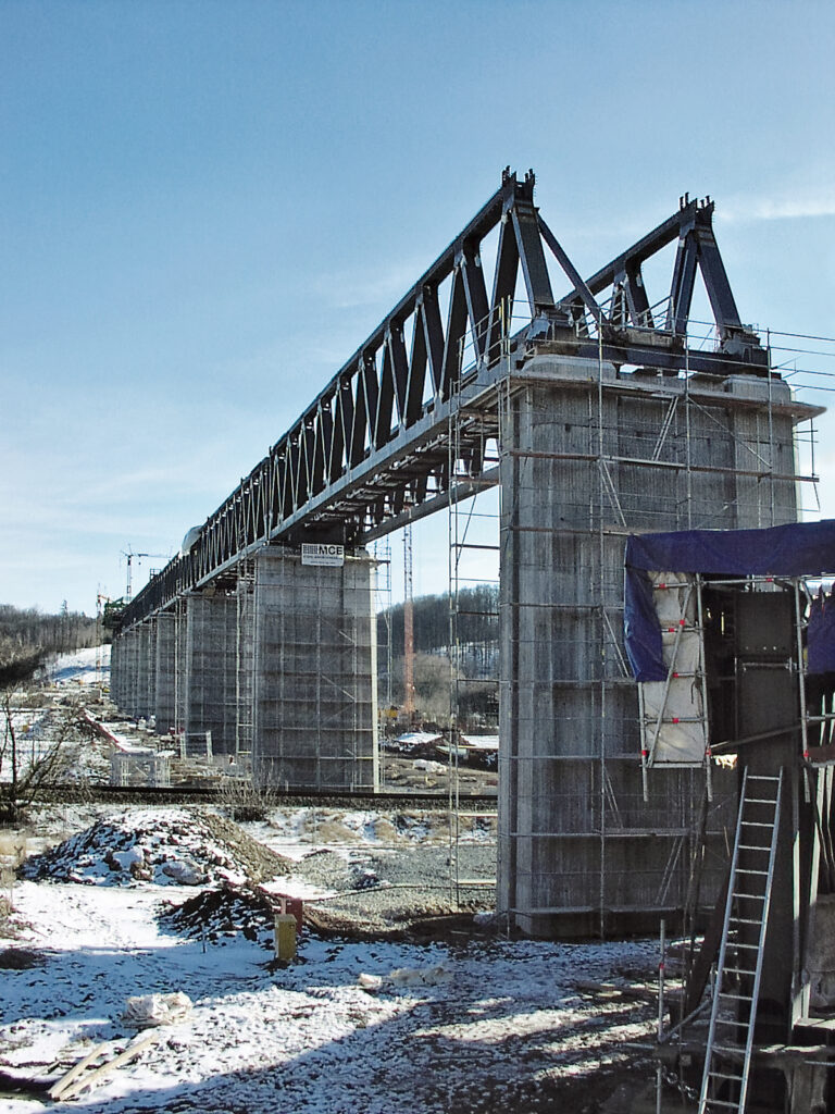 Das Bild zeigt eine große Brücke im Bau mit mehreren Betonpfeilern, die eine Stahlfachwerkkonstruktion tragen. Um die Pfeiler ist ein Gerüst aufgebaut, der Boden ist schneebedeckt und von Baumaterialien und Geräten umgeben.