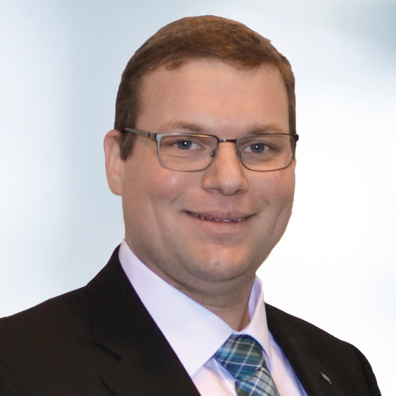 Thomas Müller trägt einen Anzug, ein weißes Hemd und eine blau karierte Krawatte. Der Hintergrund ist unscharf und zeigt ein Büro.