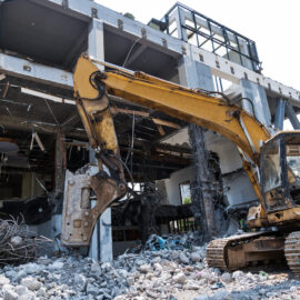 Das Bild zeigt eine Baustelle, auf der ein großer gelber Bagger ein Gebäude abreißt. Das Gebäude ist teilweise zerstört, und rundherum liegen Trümmer und Bauschutt. Der Baggerarm ist auf die Struktur gerichtet, sichtbar sind Metallverstärkungen und Betonstücke.