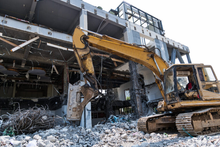 Das Bild zeigt eine Baustelle, auf der ein großer gelber Bagger ein Gebäude abreißt. Das Gebäude ist teilweise zerstört, und rundherum liegen Trümmer und Bauschutt. Der Baggerarm ist auf die Struktur gerichtet, sichtbar sind Metallverstärkungen und Betonstücke.