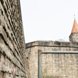 Das Bild zeigt einen Abschnitt der Nürnberger Stadtmauer aus Sandstein. Im Vordergrund ist die strukturierte Oberfläche der Mauer zu sehen, im Hintergrund ein Turm mit rotem Ziegeldach sowie weitere Teile der Befestigungsanlage. In der Bildmitte ist ein Metallgeländer erkennbar.