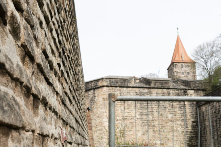 Das Bild zeigt einen Abschnitt der Nürnberger Stadtmauer aus Sandstein. Im Vordergrund ist die strukturierte Oberfläche der Mauer zu sehen, im Hintergrund ein Turm mit rotem Ziegeldach sowie weitere Teile der Befestigungsanlage. In der Bildmitte ist ein Metallgeländer erkennbar.