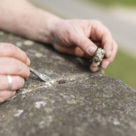Das Bild zeigt eine Nahaufnahme von zwei Händen, die an einer Sandsteinmauer arbeiten, der Nürnberger Stadtmauer. Eine Hand hält ein kleines Meißelwerkzeug, die andere ein herausgelöstes Stück Stein oder Material. Der Fokus liegt auf der Entnahme einer Probe aus dem Sandstein.