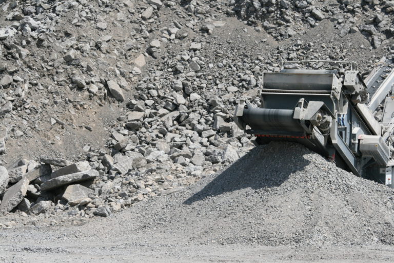 Das Bild zeigt eine Gesteinsbrechmaschine im Einsatz in einem Steinbruch. Große Steine werden zu kleineren, kiesartigen Stücken zerkleinert und über ein Förderband in einem Haufen abgelegt. Im Hintergrund ist felsiges Gelände mit unterschiedlich großen Steinen zu sehen.