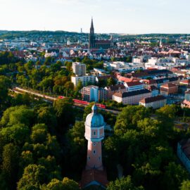 Luftaufnahme von Neu-Ulm mit einem markanten Turm mit blauer Kuppel im Vordergrund, umgeben von Bäumen; in der Mitte fährt ein roter Zug durch die Stadt, im Hintergrund ist das Ulmer Münster deutlich sichtbar vor der städtischen Kulisse.