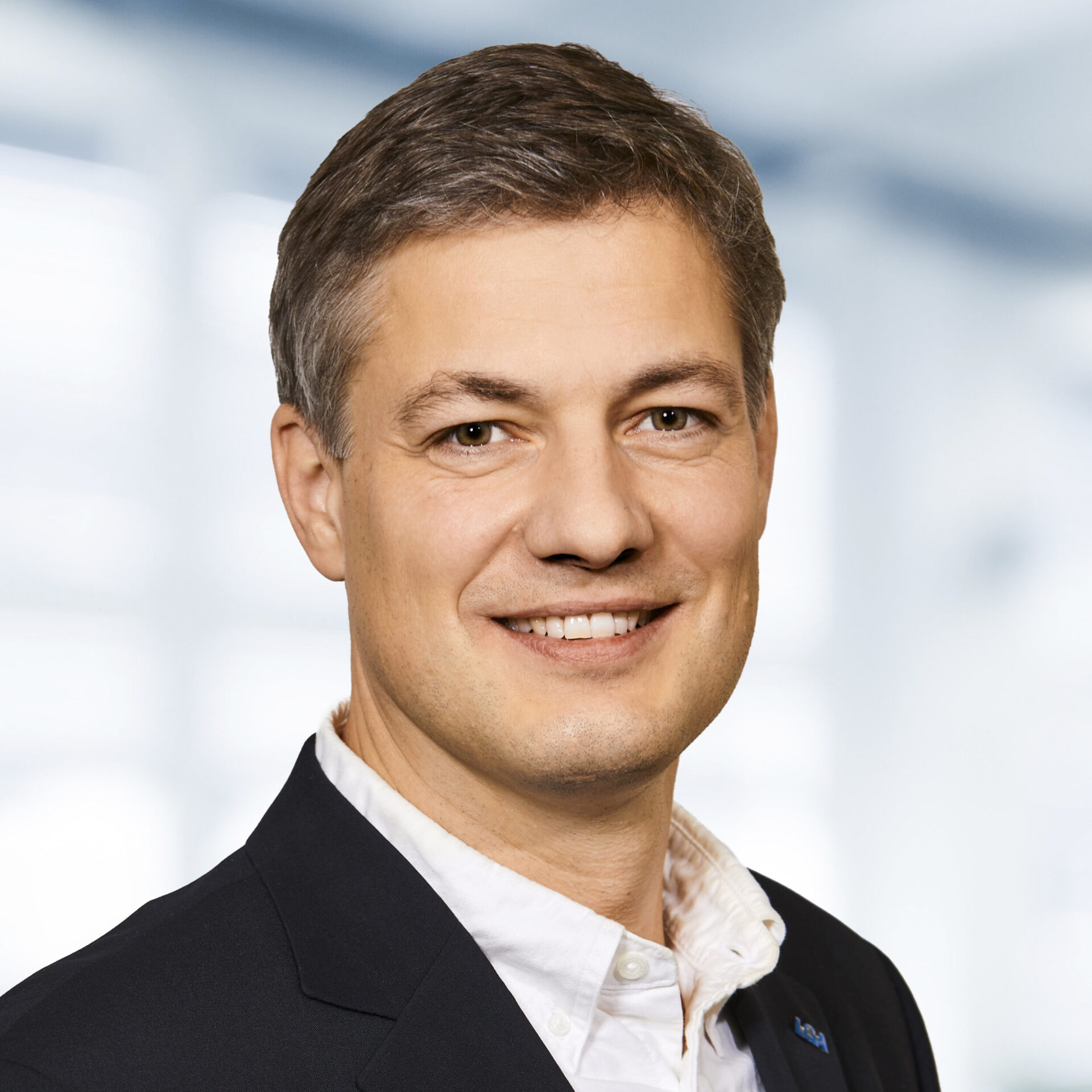 Nikolas Nissen trägt einen dunklen Blazer und ein weißes Hemd. Der Hintergrund ist unscharf und zeigt ein Büro.