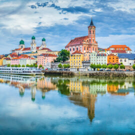 Panoramablick auf Passau mit bunten Gebäuden entlang des Flussufers, darunter der markante Dom St. Stephan mit grünen Kuppeln; die ruhige Wasseroberfläche spiegelt die Stadt und den Himmel wider und erzeugt ein malerisches Gesamtbild.
