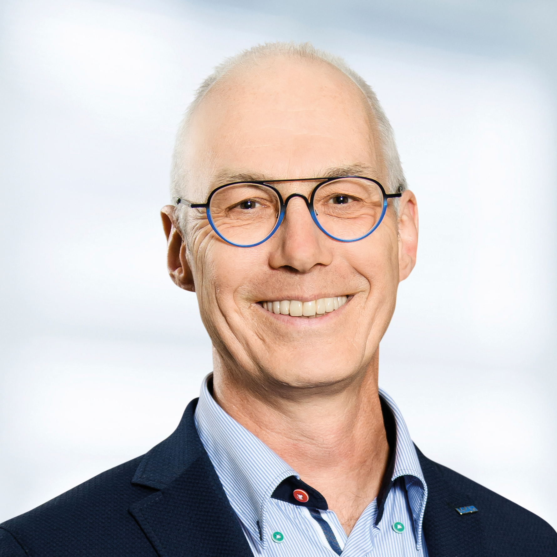 Günter Peters trägt eine Brille, ein blaues Sakko und ein hellblaues Hemd mit Knöpfen. Der Hintergrund ist unscharf und zeigt ein Büro.