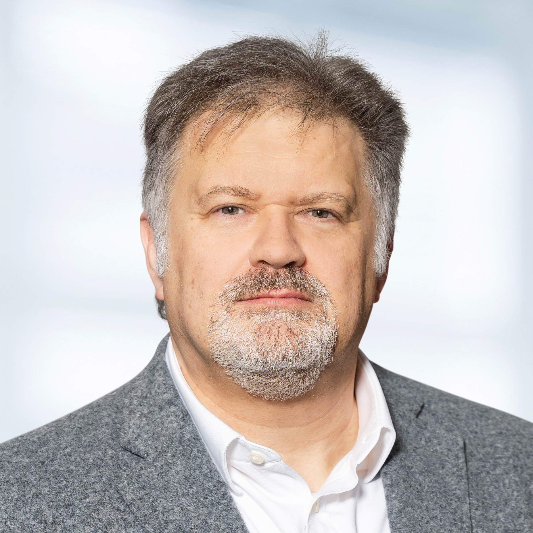 Rolf Plechatz trägt ein graues Sakko und ein weißes Hemd. Der Hintergrund ist unscharf und zeigt ein Büro.