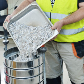 Das Bild zeigt eine Person mit gelber Sicherheitsweste und beigefarbener Hose, die in einem Labor- oder Industriebereich zerkleinerte Steine oder Kies aus einem Metallbehälter in ein Siebgerät schüttet. Die Siebe sind übereinandergestapelt und dienen vermutlich der Materialanalyse.