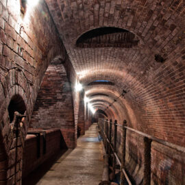 Das Bild zeigt einen unterirdischen Tunnel mit gemauerten Wänden und einem gewölbten Deckengewölbe. Der Tunnel ist gut beleuchtet, mit mehreren Wandlampen, die einen Weg in der Mitte ausleuchten. Auf einer Seite verläuft ein Metallgeländer, und im Tunnel sind verschiedene Rohre und bauliche Elemente sichtbar.