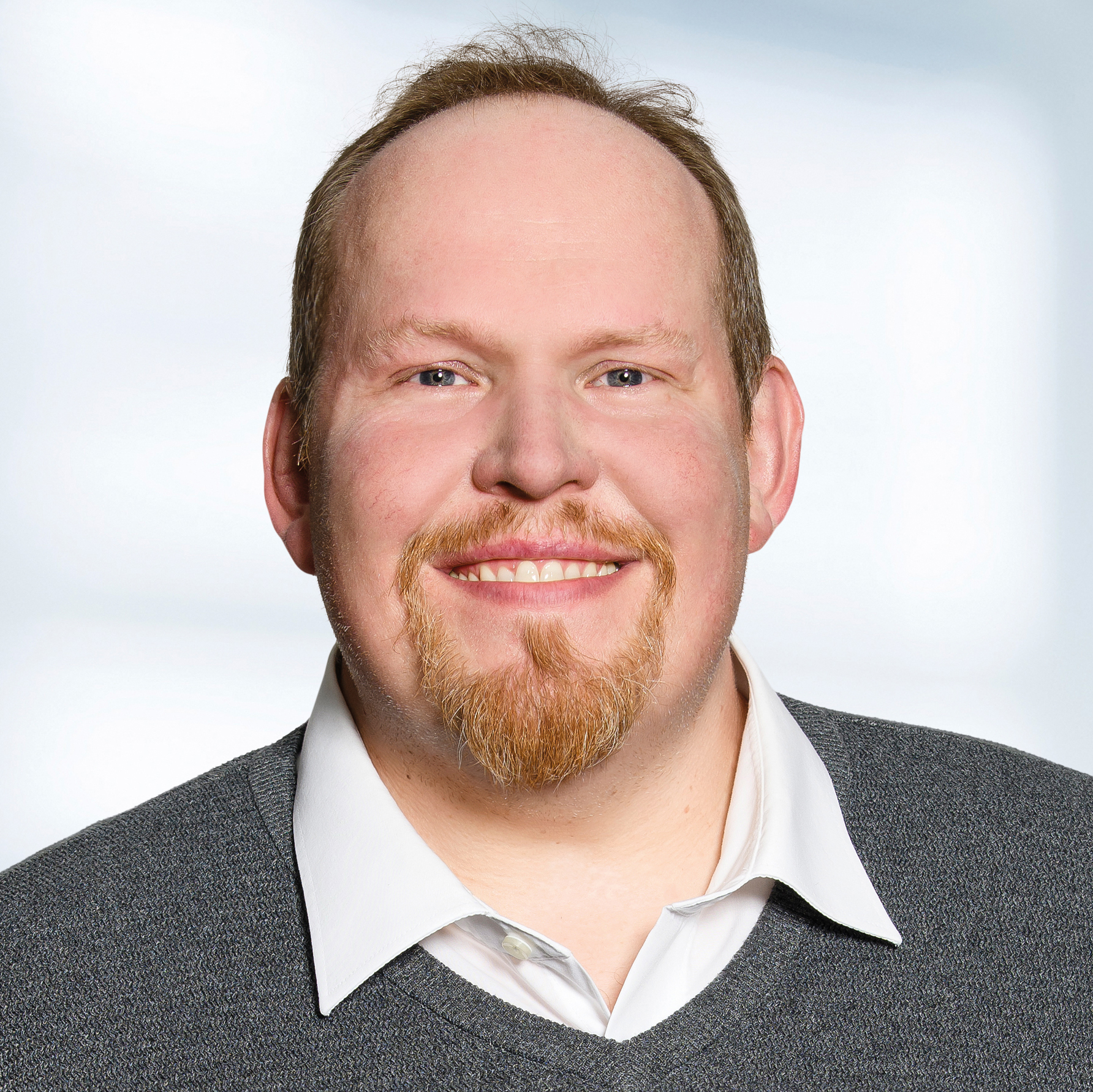 Michael Schäfer trägt ein weißes Hemd mit Kragen unter einem grauen Pullover. Der Hintergrund ist unscharf und zeigt ein Büro.