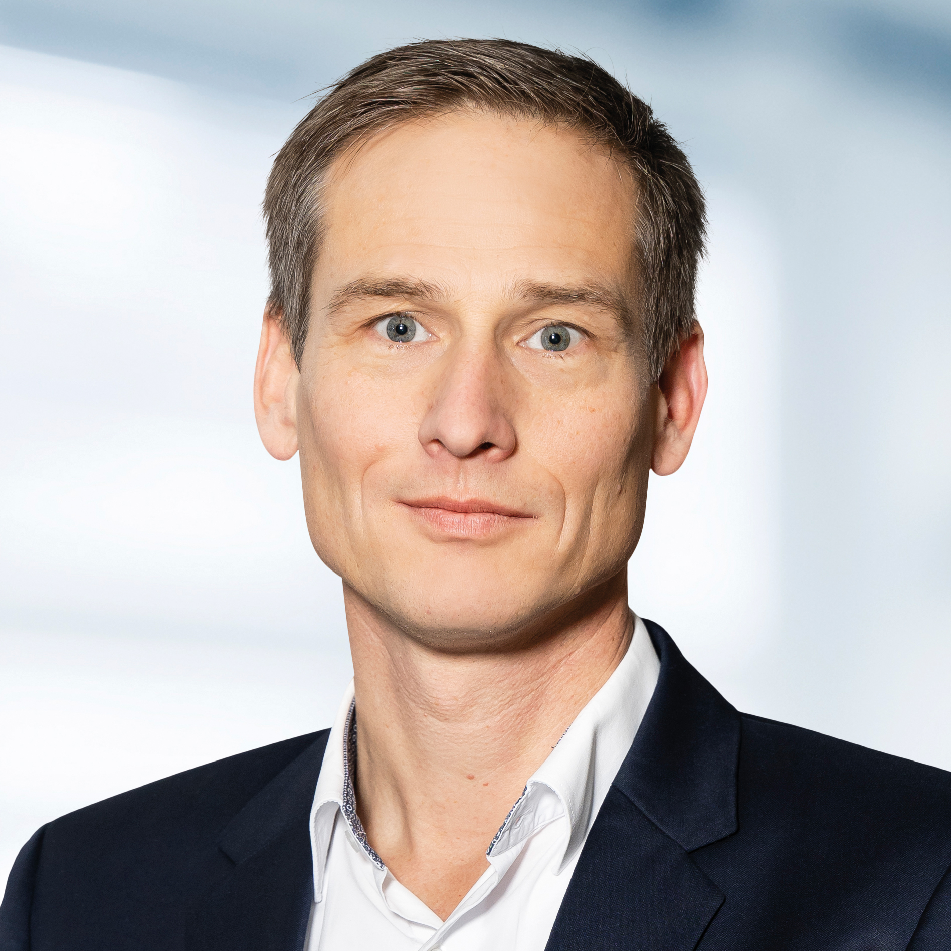 Dr. Jörg Schaffrath trägt ein dunkles Sakko und ein weißes Hemd mit Kragen. Der Hintergrund ist unscharf und zeigt ein Büro.
