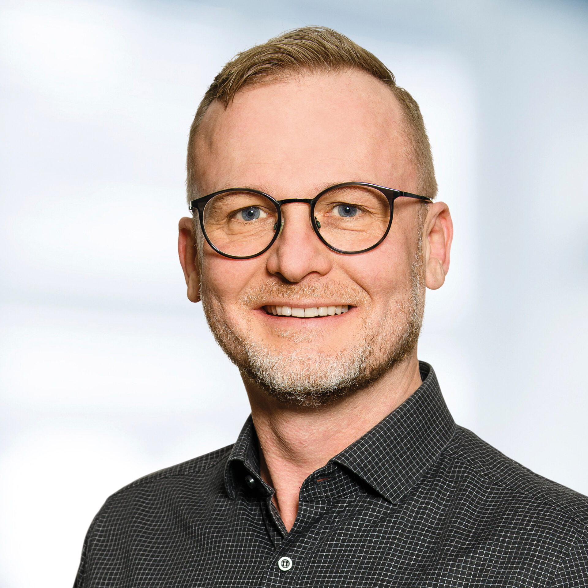 Mark Schüßler trägt eine Brille und ein dunkel kariertes Hemd. Der Hintergrund ist unscharf und zeigt ein Büro.