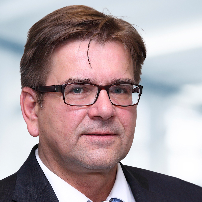 Ulrich Sieler trägt eine Brille und einen Anzug. Der Hintergrund ist unscharf und zeigt ein Büro.