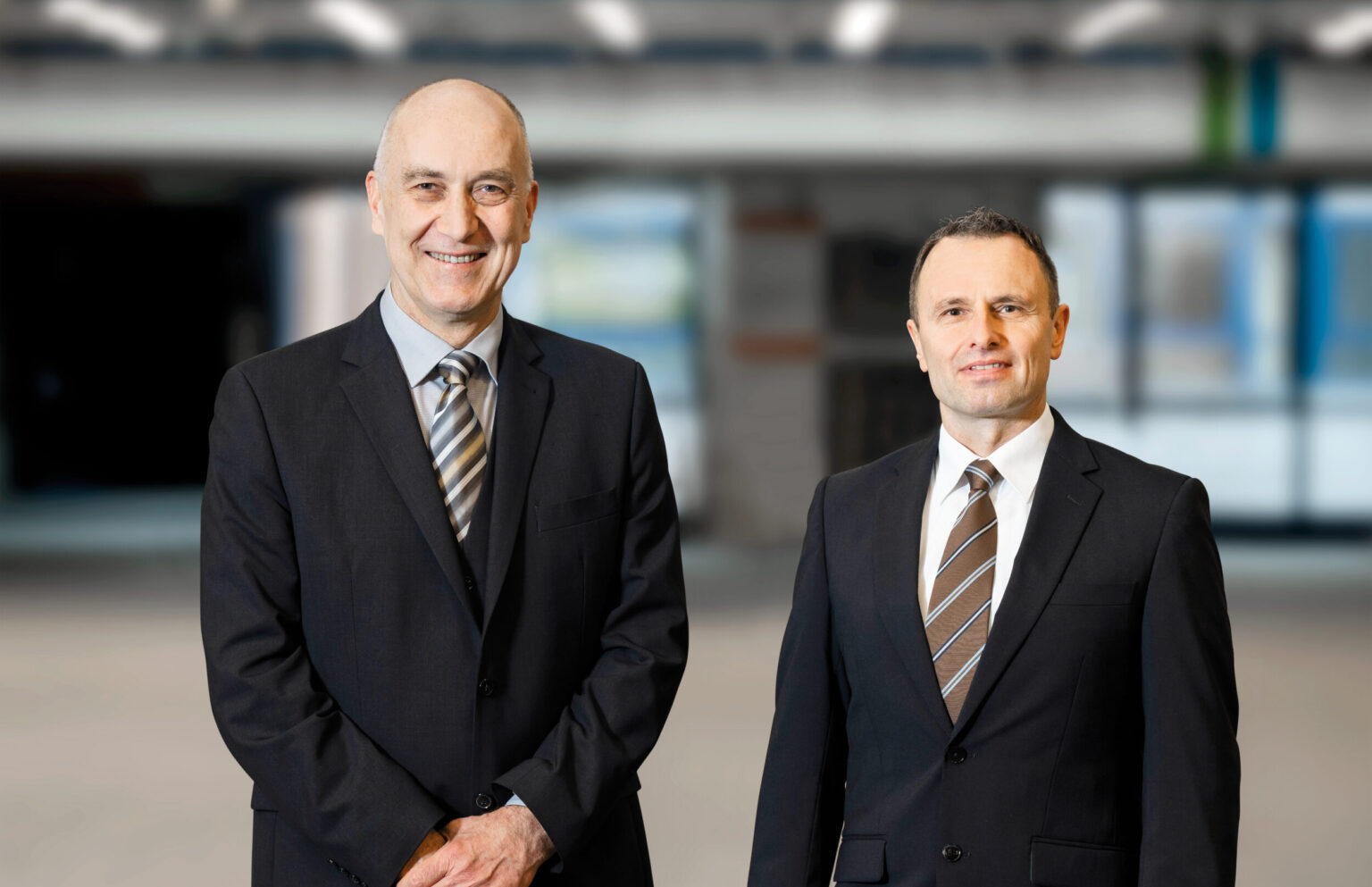 Zwei Personen in dunklen Anzügen mit weißen Hemden und gestreiften Krawatten stehen in einem Foyer. Der Hintergund ist stark verschwommen.