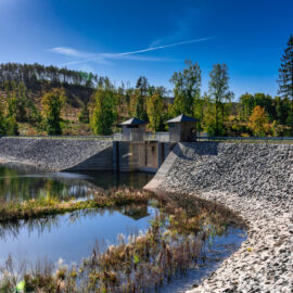 Das Bild zeigt ein Ausgleichsbecken mit einer Staumauer aus Beton. Auf der Mauer befinden sich zwei kleine Gebäude, zur Steuerung der Anlage. Das Becken ist von steinernen Uferbefestigungen umgeben, im Hintergrund sind Bäume und Hügel unter einem klaren blauen Himmel zu sehen.