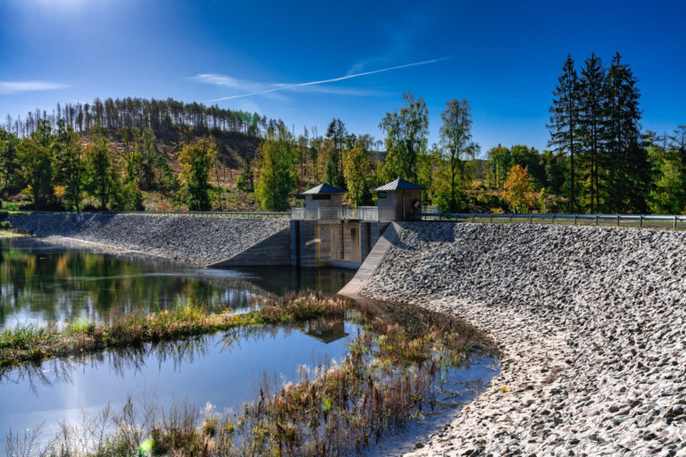 Das Bild zeigt ein Ausgleichsbecken mit einer Staumauer aus Beton. Auf der Mauer befinden sich zwei kleine Gebäude, zur Steuerung der Anlage. Das Becken ist von steinernen Uferbefestigungen umgeben, im Hintergrund sind Bäume und Hügel unter einem klaren blauen Himmel zu sehen.