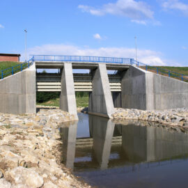 Das Bild zeigt eine Betonbrücke über einem kleinen Gewässer. Die Brücke hat drei große rechteckige Wasserdurchlässe, die von massiven Betonpfeilern getragen werden. Sie dienen als Staudamm. Entlang der Brücke verläuft ein blaues Metallgeländer, die Ufer sind mit Steinen gegen Erosion gesichert.