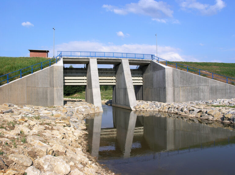 Das Bild zeigt eine Betonbrücke über einem kleinen Gewässer. Die Brücke hat drei große rechteckige Wasserdurchlässe, die von massiven Betonpfeilern getragen werden. Sie dienen als Staudamm. Entlang der Brücke verläuft ein blaues Metallgeländer, die Ufer sind mit Steinen gegen Erosion gesichert.