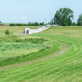 Das Bild zeigt eine grüne Landschaft mit einer Hochwasserschutzanlage. Die Anlage besteht aus einer Betonmauer und einem Tor, das auf einer erhöhten Fläche liegt. Ein geschwungener Weg führt zum Staudamm, umgeben von Wiesen und Bäumen im Hintergrund.